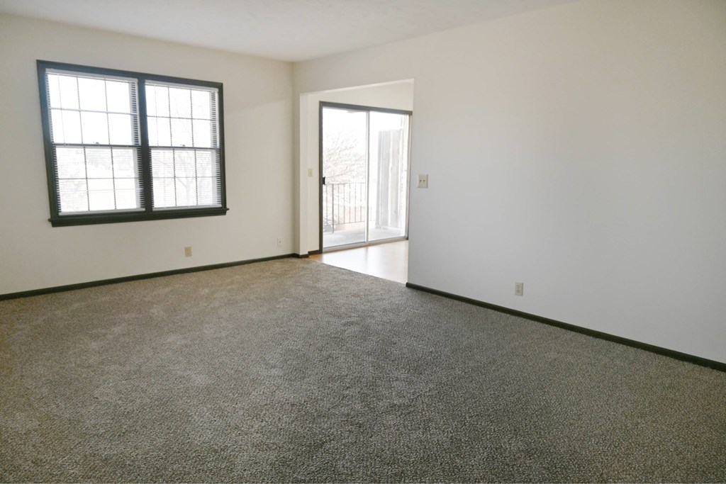 A room with a carpeted floor, a window with blinds, and a door leading to another room.