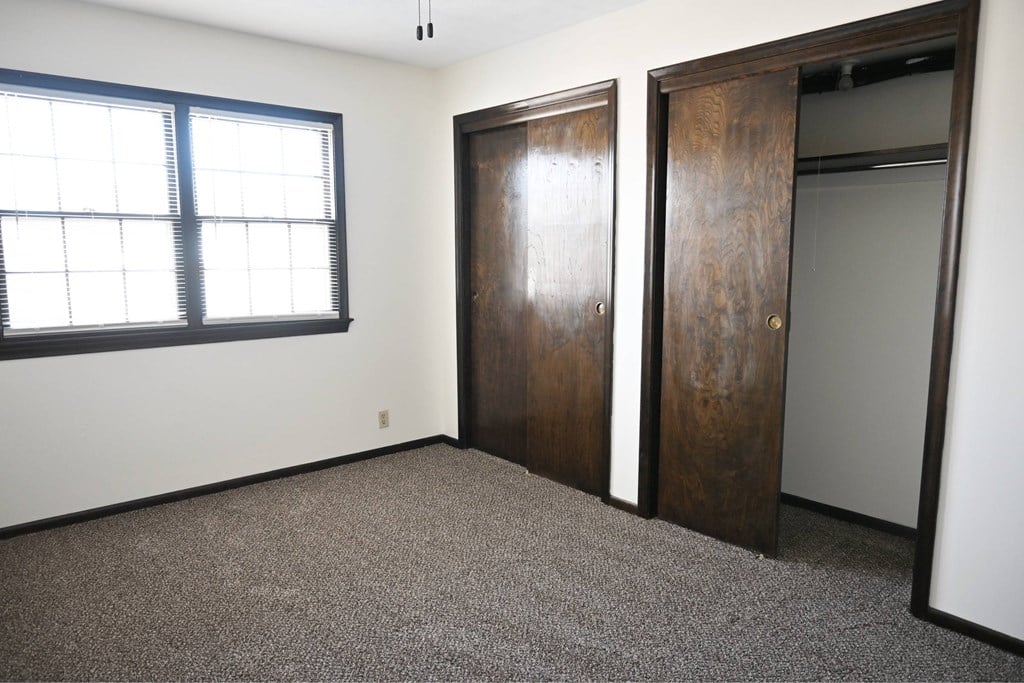 A room with a carpeted floor, a window with blinds, and two wooden doors.