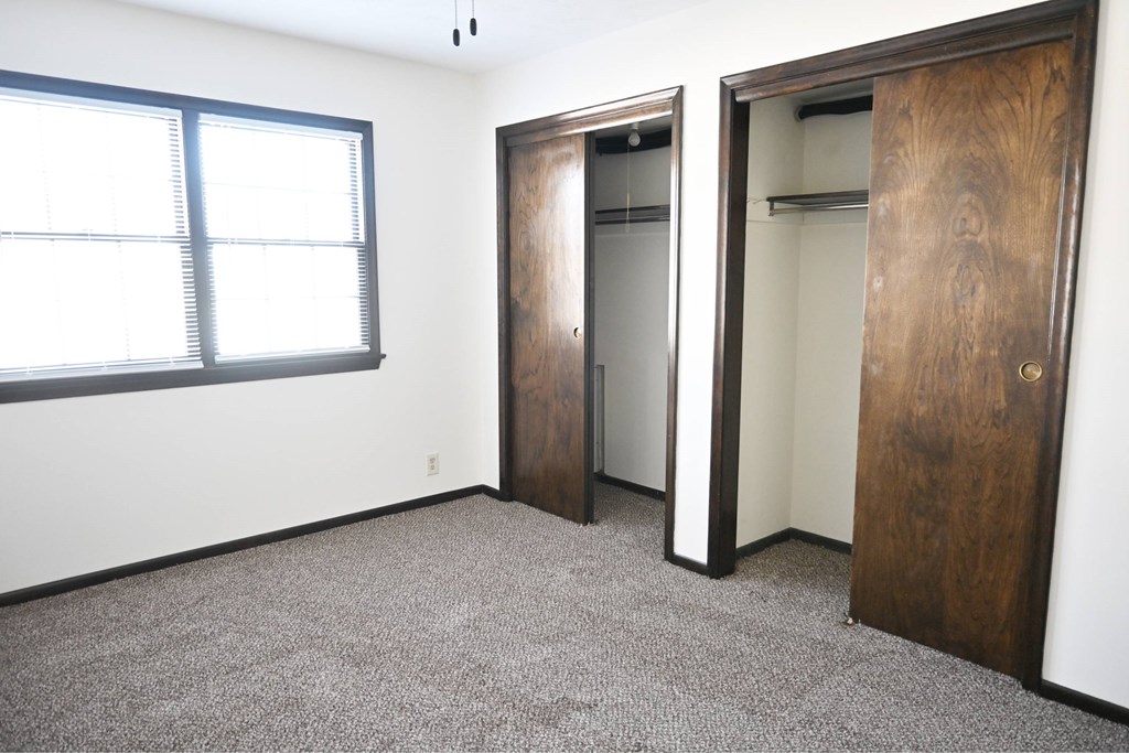 A room with a carpeted floor, a window with blinds, and two wooden doors.