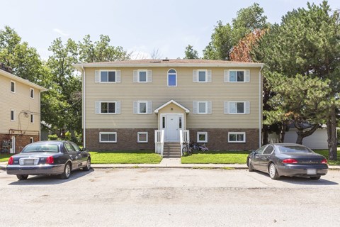 A two-story building with a beige facade and a white door is surrounded by trees and has two cars parked in front.