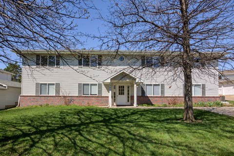 A house with a grey roof and a white door is surrounded by a green lawn.