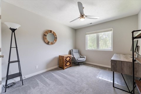 A room with a grey carpet, a ceiling fan, a window, and a wooden box on the floor.