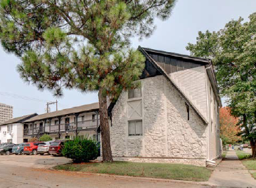 A small stone building sits in front of a large building and a tree.