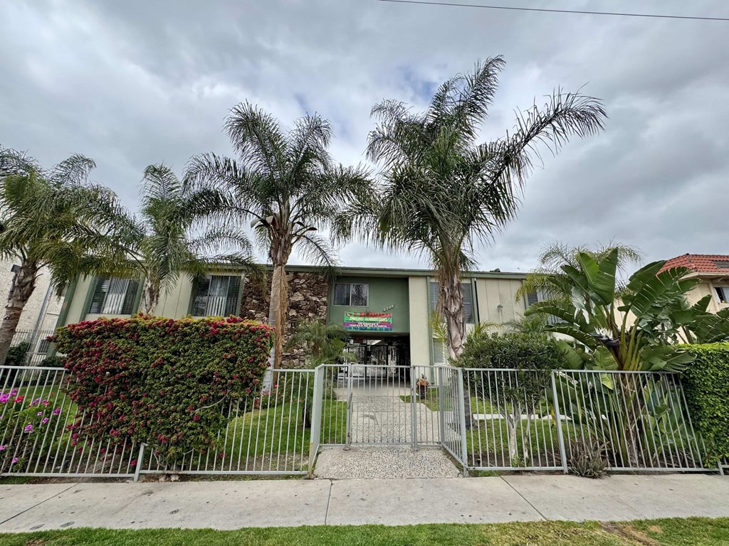 A building with a green awning is surrounded by a metal fence and palm trees.