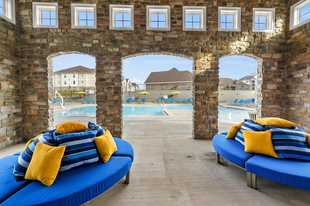 A blue cushioned bench with yellow pillows is in the foreground of a stone archway with a pool and buildings in the background.