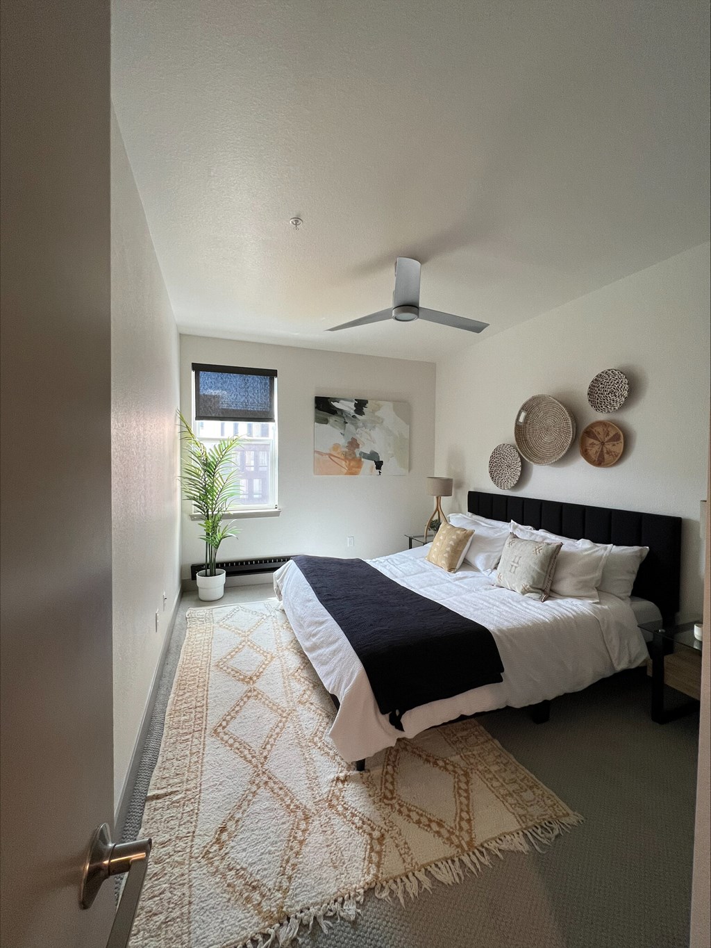 A bedroom with a bed, a rug, a ceiling fan, and a plant.