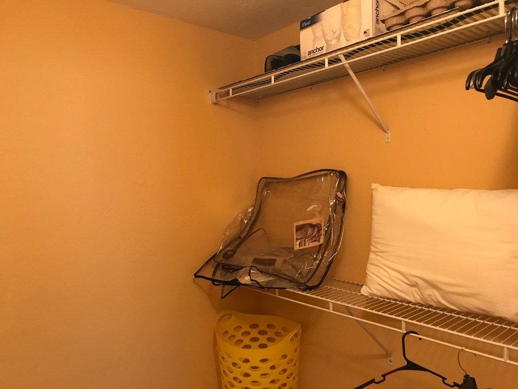 A yellow laundry basket sits on a shelf under a white pillow.
