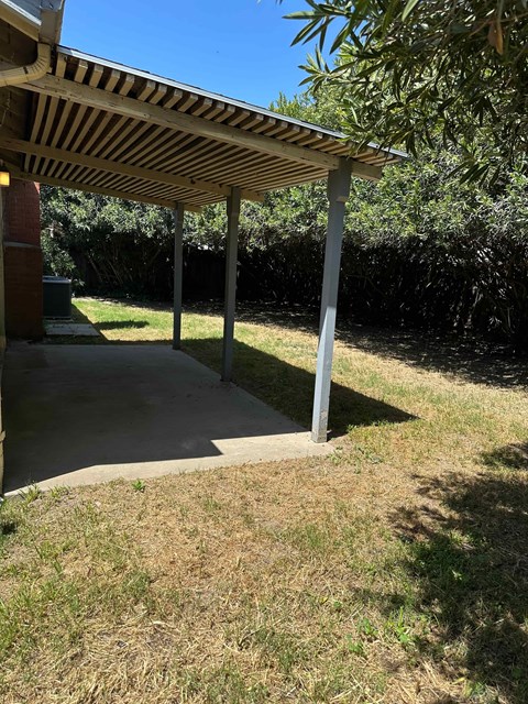 A patio with a roof and pillars is surrounded by grass and trees.