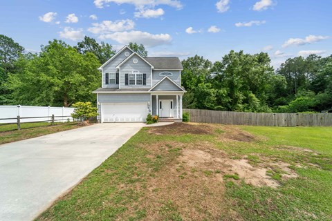 A house with a grey roof and white walls is surrounded by a grassy area and a wooden fence.