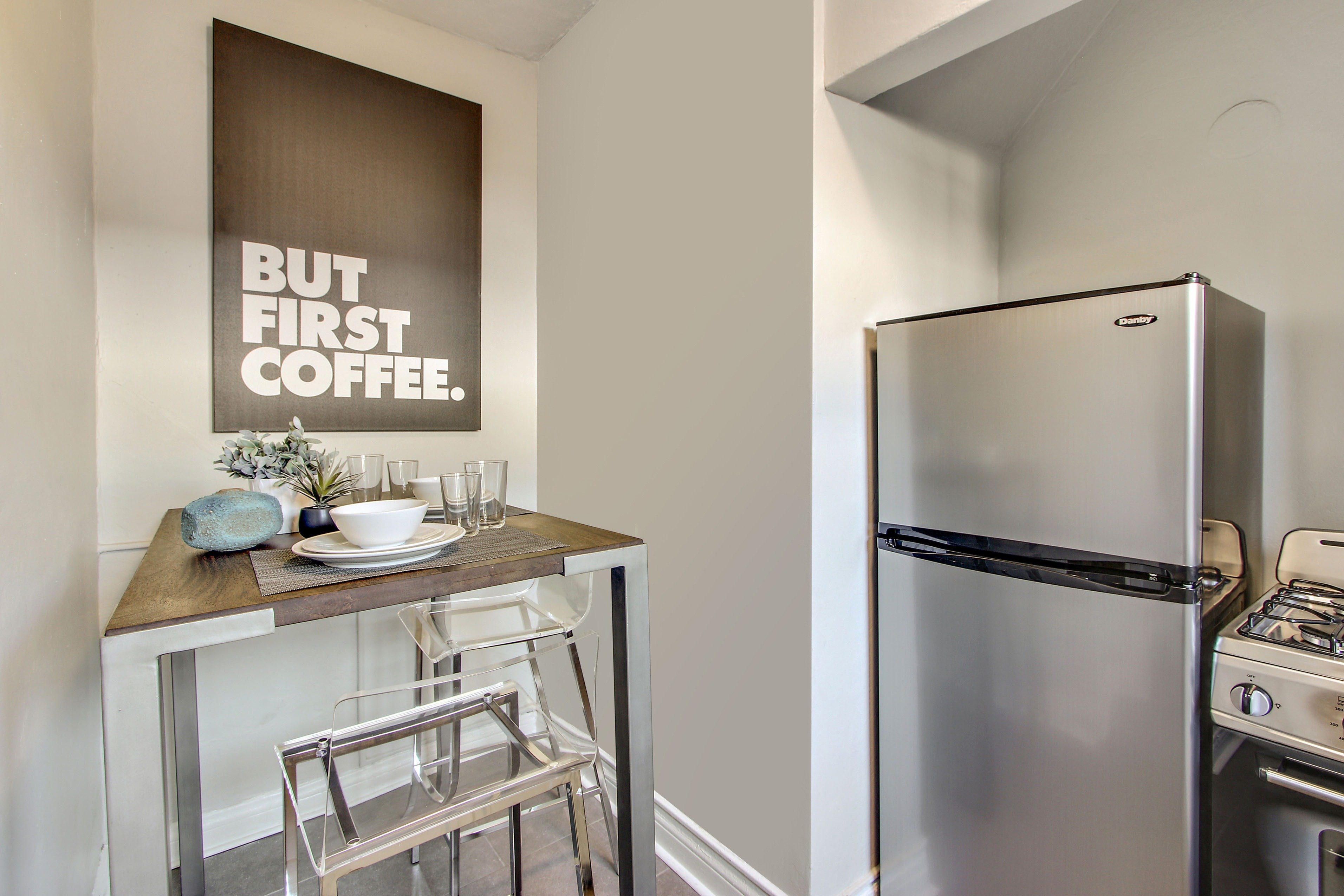A kitchen with a stainless steel refrigerator and a table with a sign that says "but first coffee.".