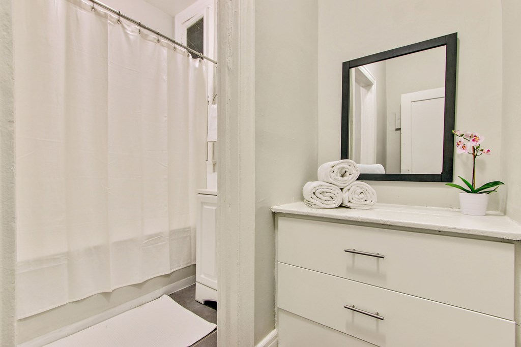 A bathroom with a white shower curtain, a white countertop with a mirror, two towels, and a potted plant.