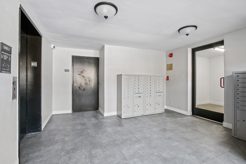 A room with a grey floor and white walls has a fire extinguisher on the wall and a metal door on the left.