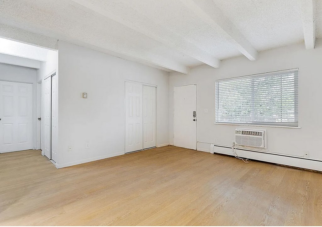 A room with white walls and wooden flooring, with a window covered by blinds.