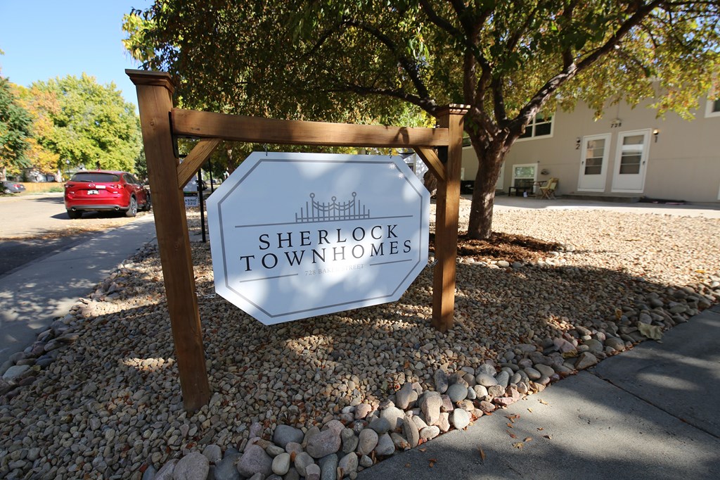 A sign that says Sherlock Townhomes stands in front of a tree and a building.
