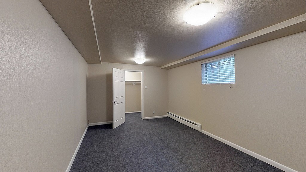 A room with a carpeted floor, a window with blinds, and a door leading to another room.
