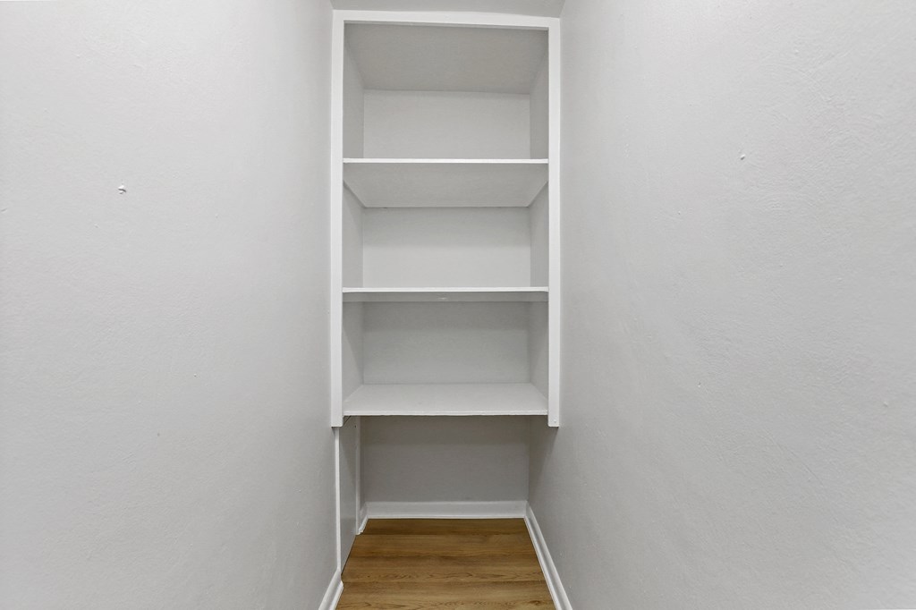 A white shelving unit with four shelves is in a corner of a room with white walls.