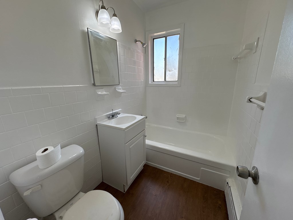 A white toilet with a roll of toilet paper on top sits in a bathroom with white tiles.