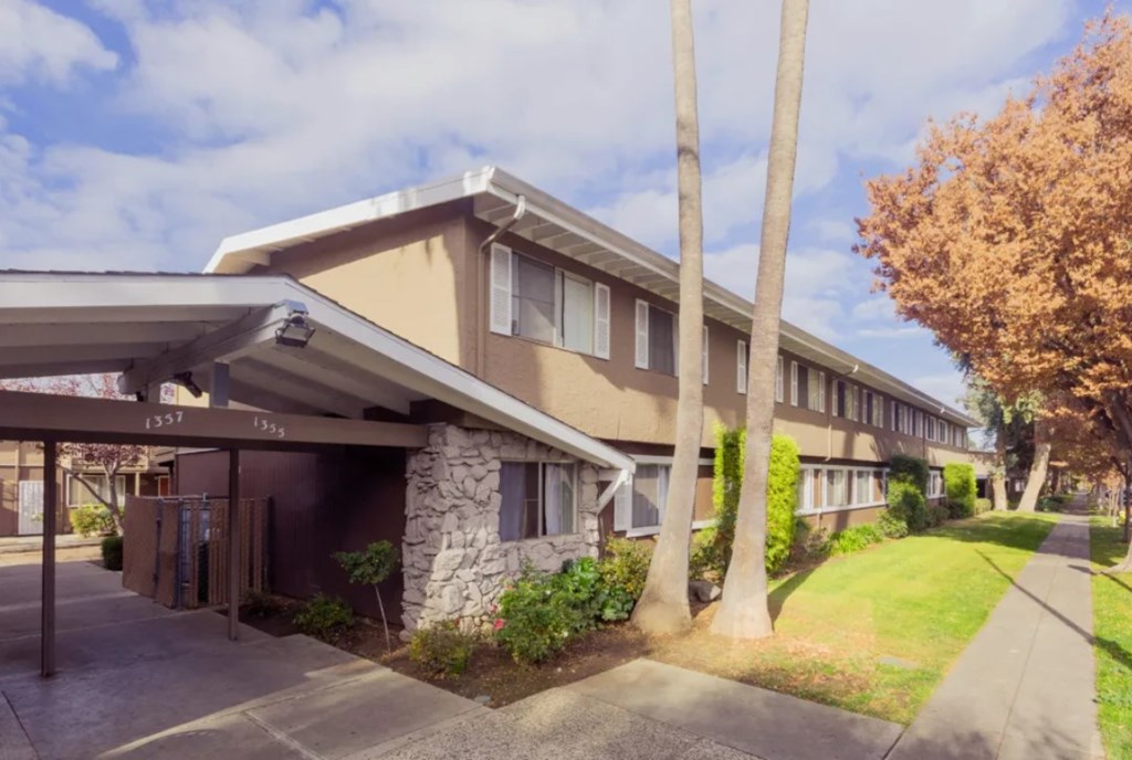 A building with a brown and beige exterior is surrounded by trees and a stone pillar.