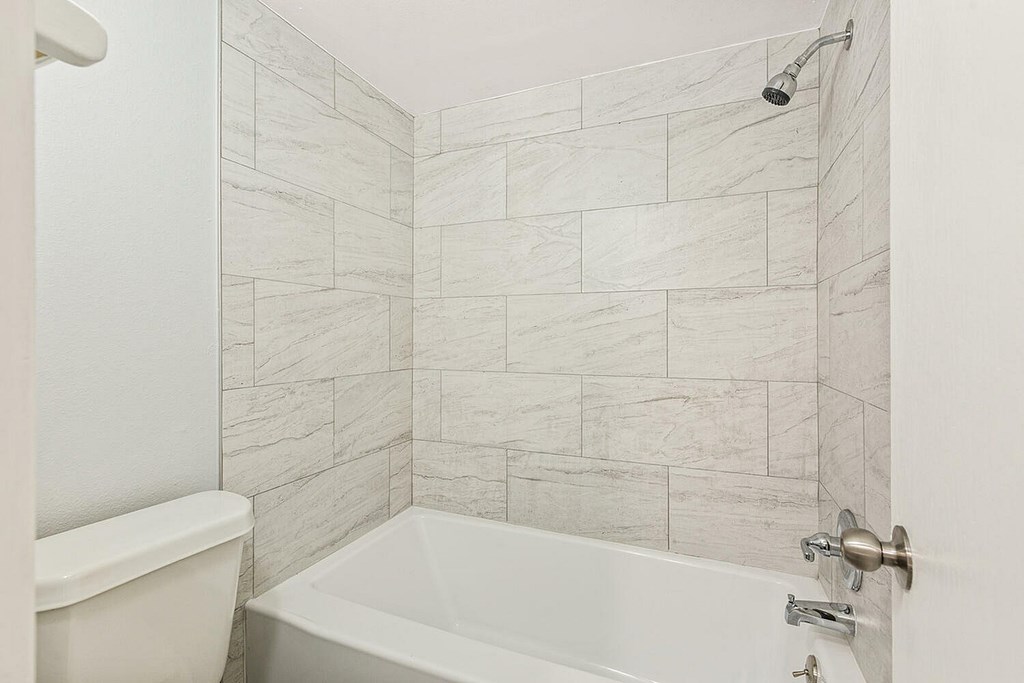 A white bathtub sits next to a white toilet in a bathroom with a marble tile wall.