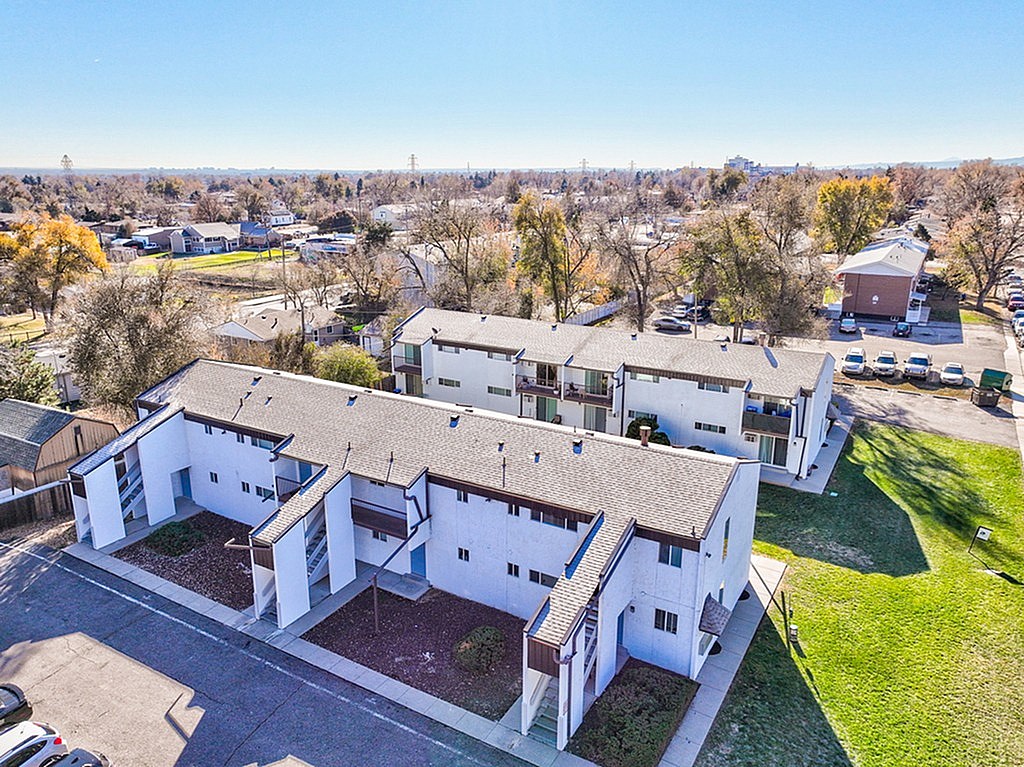 A large white building with a brown roof is surrounded by a grassy area and other buildings in the background.