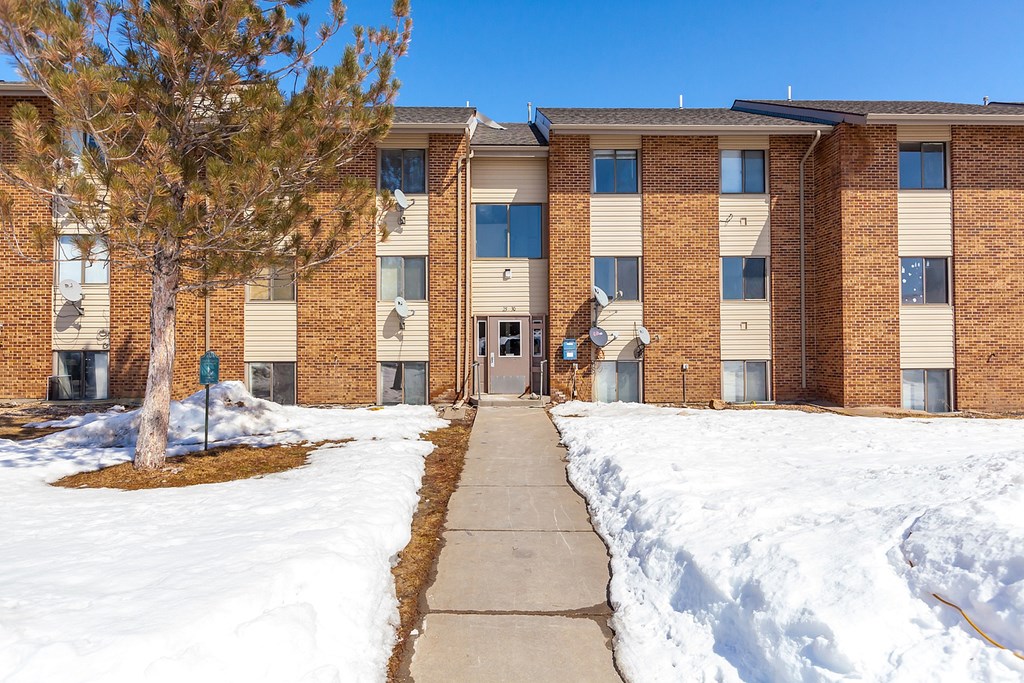 A snow-covered walkway leads to a brick apartment building with a green sign on the left.