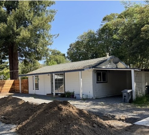 A house with a white exterior and a grey roof is surrounded by trees and has a pile of dirt in front of it.