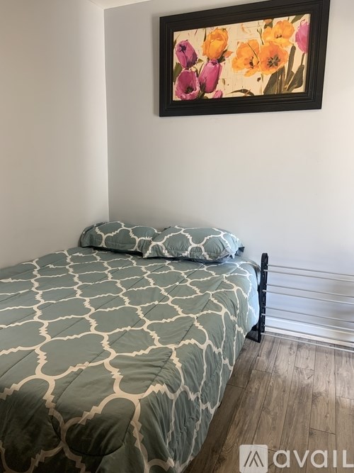 A bed with a patterned cover and a pillow is in the foreground with a painting of flowers on the wall behind it.