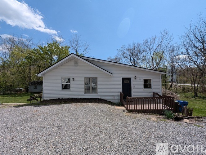 A white house with a black door and windows is surrounded by a gravel driveway.