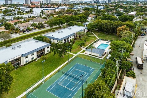 A tennis court is surrounded by a grassy area and apartment buildings.