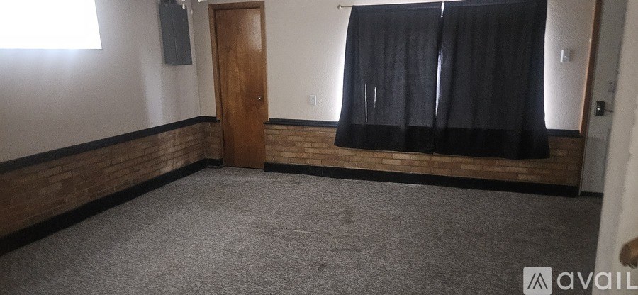 A room with a carpeted floor, a wooden door, and a window with black curtains.