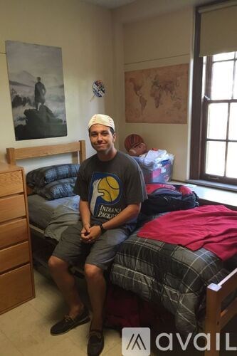 A man in a black shirt and plaid shorts sits on a bed in a room with a world map on the wall.