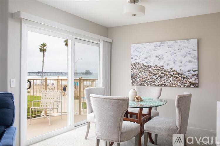 A living room with a glass table and chairs, a painting, and a view of a balcony with a palm tree.