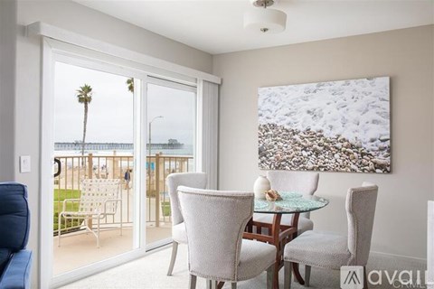 A living room with a glass table and chairs, a painting, and a view of a balcony with a palm tree.