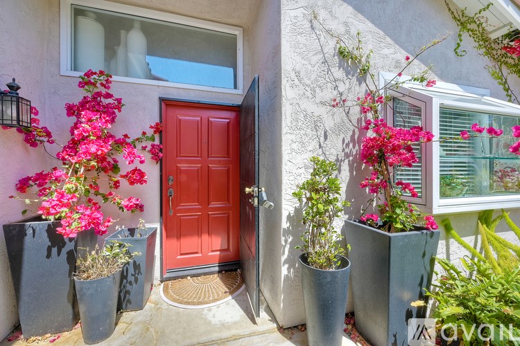 A red door is flanked by two grey planters with pink flowers.