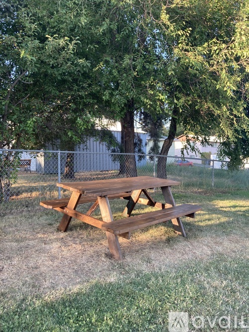 A wooden picnic table sits in a grassy area with trees and a fence in the background.