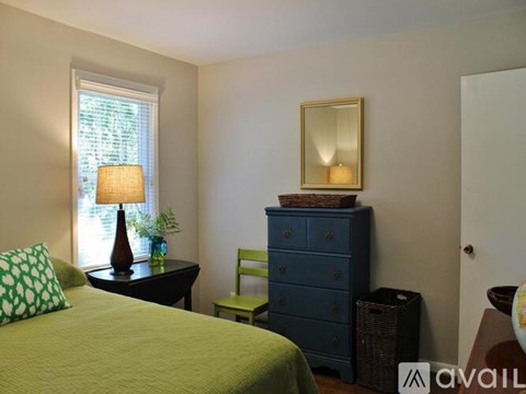 A bedroom with a bed, a dresser, a chair, a table with a lamp and a mirror.