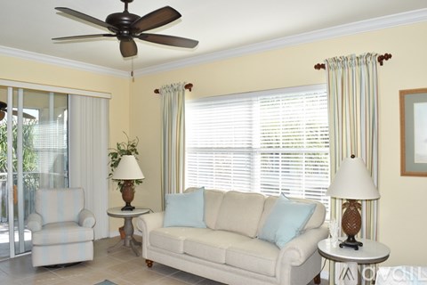 A living room with a white couch, a fan, and a table with a lamp on it.