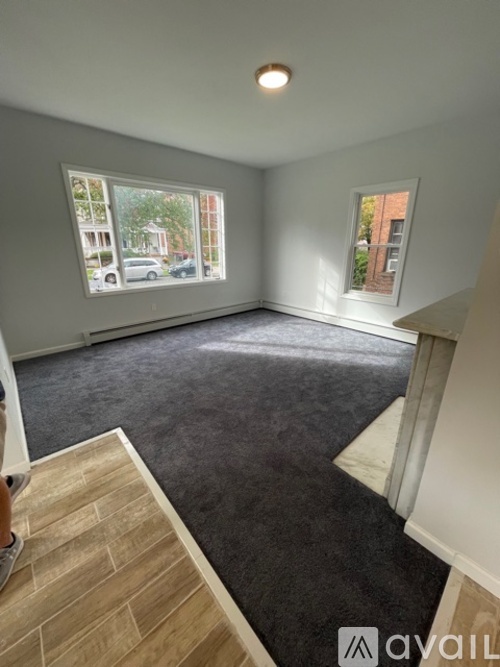A room with a carpeted floor, a window with a view of a building and trees, and a wooden staircase leading to another room.