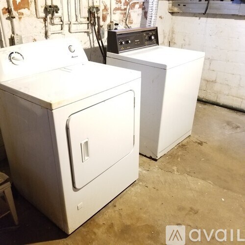 Two white front loading washing machines in a room with a white wall and pipes.