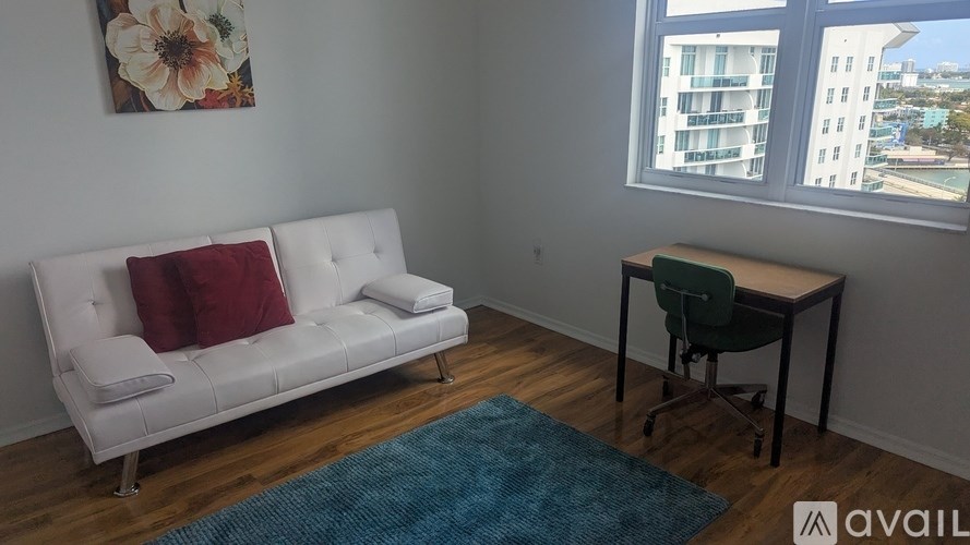 A white couch with red pillows sits in a room with a blue rug and a window showing a cityscape.