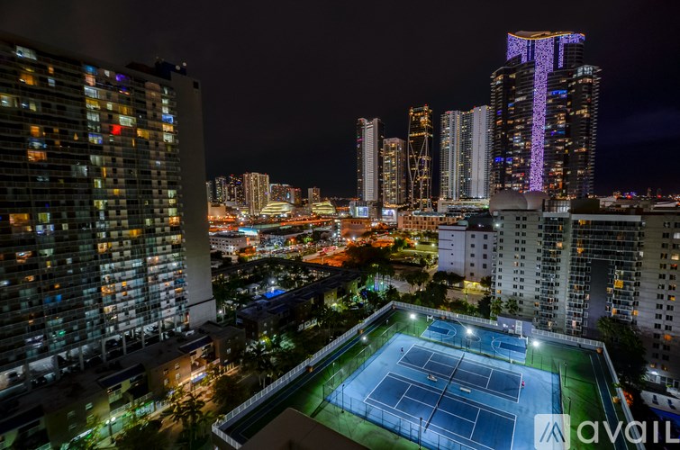 A tennis court is lit up at night in a city with tall buildings.