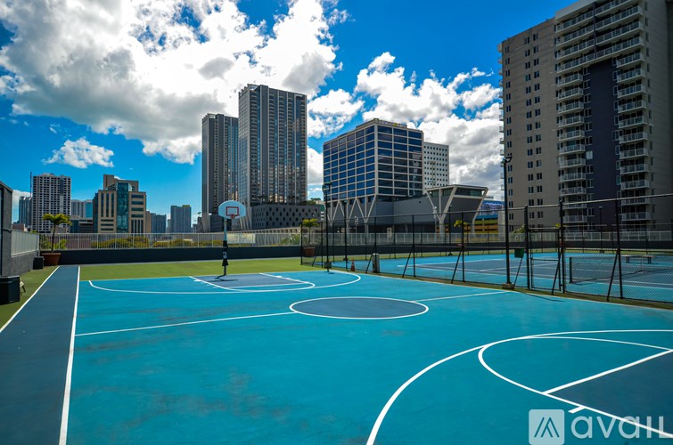 A basketball court with a blue surface and white lines is surrounded by tall buildings under a blue sky with clouds.