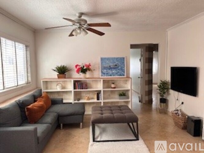 A living room with a grey couch, a brown ottoman, a white bookshelf, a ceiling fan, and a flat screen TV.