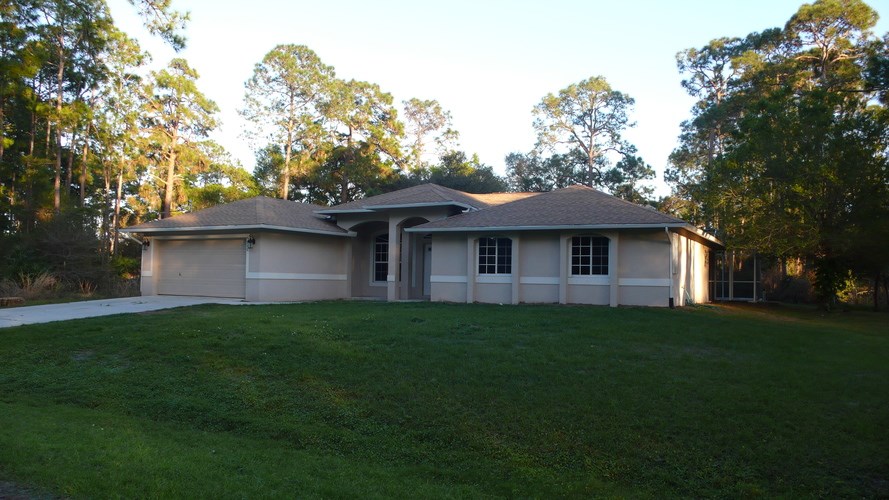A house with a grey roof and white walls is surrounded by green grass and trees.