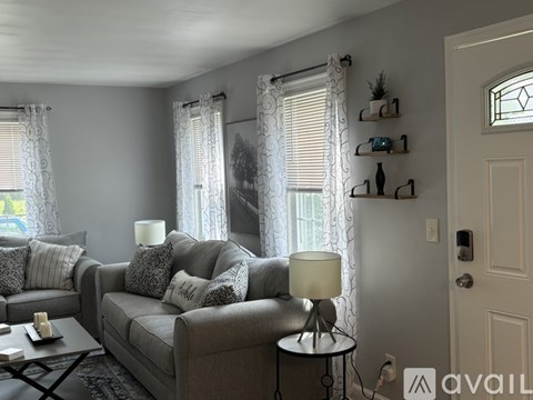 A living room with a grey couch, a table with a lamp, and a wall with shelves.