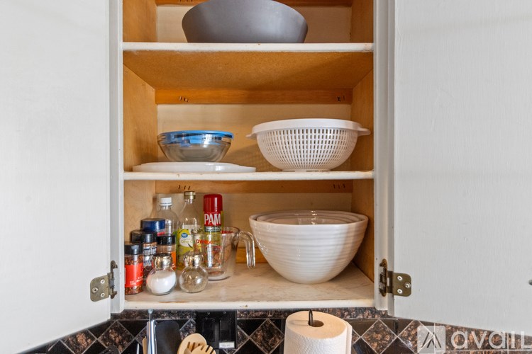 A kitchen cabinet is open, revealing a bowl, a colander, and various spices and containers.