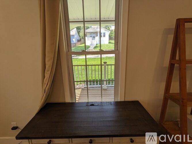 A wooden table is in the foreground of a room with a view of a house and a fence outside.