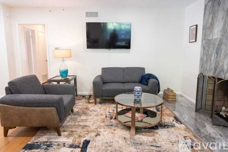 A living room with a grey couch, a grey armchair, a grey chair, a coffee table, a rug, a fireplace, a painting, a lamp, and a TV.