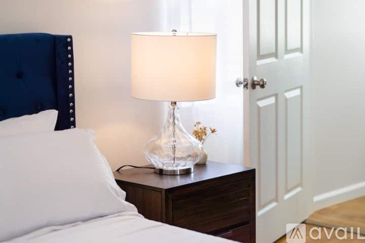 A bedroom with a white bed, a blue headboard, a nightstand with a lamp and a vase with flowers.