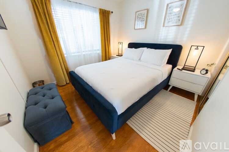 A bedroom with a large bed, a small bedside table, and a window with yellow curtains.